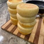 Thick homemade English muffins with golden tops stacked on a wooden cutting board beside a skillet.