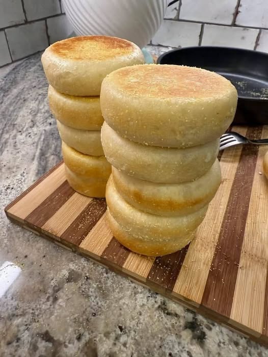Thick homemade English muffins with golden tops stacked on a wooden cutting board beside a skillet.