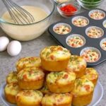 Golden savory muffins with diced red vegetables and herbs stacked on a plate beside a filled muffin pan.