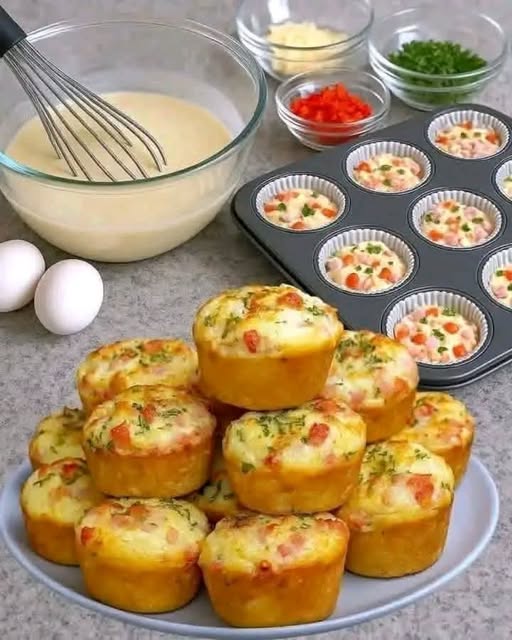 Golden savory muffins with diced red vegetables and herbs stacked on a plate beside a filled muffin pan.