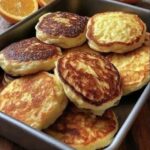 Thick golden-brown pancakes arranged in a metal tray with fresh orange halves behind them.