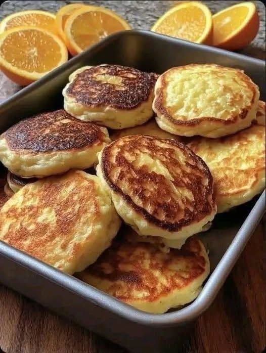 Thick golden-brown pancakes arranged in a metal tray with fresh orange halves behind them.