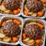 Four individual aluminum trays holding browned beef patties over sliced potatoes and carrots.