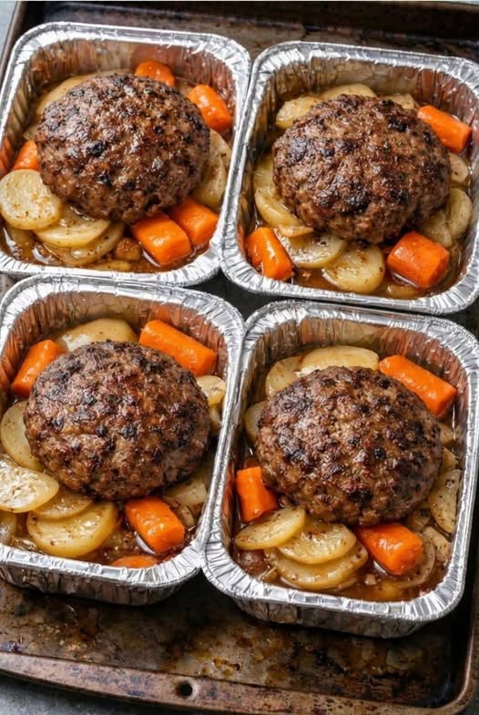 Four individual aluminum trays holding browned beef patties over sliced potatoes and carrots.