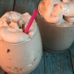 Thick homemade keto chocolate frosty in a clear glass with a spoon on a rustic wooden table.