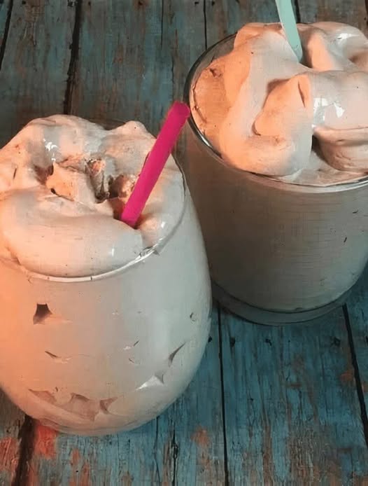 Thick homemade keto chocolate frosty in a clear glass with a spoon on a rustic wooden table.