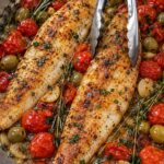Golden seared white fish fillets in a skillet with cherry tomatoes, green olives, garlic cloves, rosemary, and thyme.